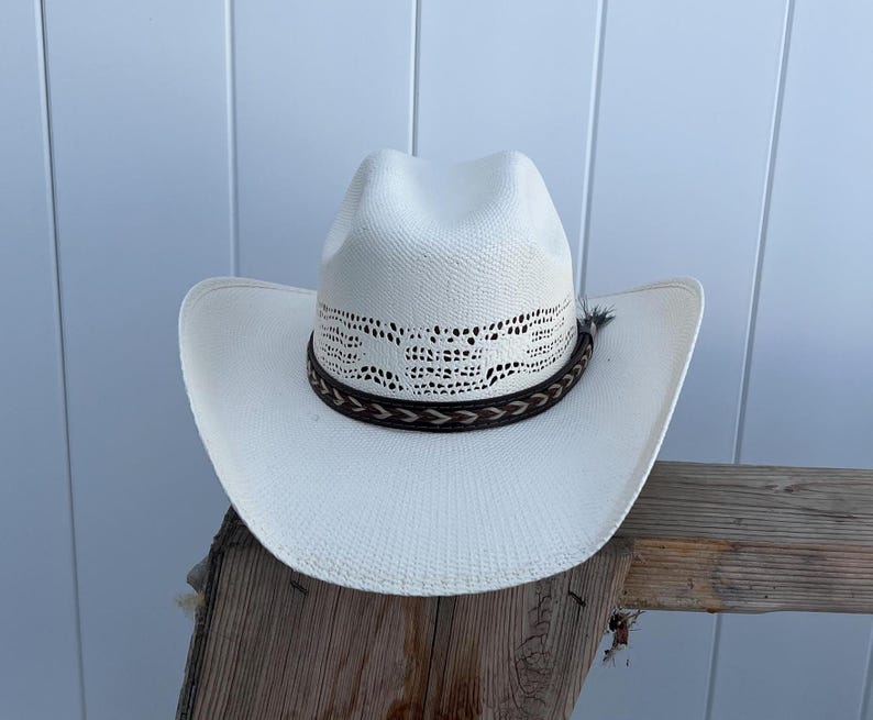 May include: A white straw cowboy hat with a brown leather band and a white lace design. The hat is sitting on a wooden surface.