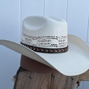 May include: A white straw cowboy hat with a brown leather band and a woven design around the crown.