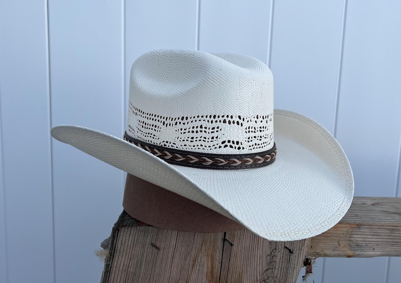 May include: A white straw cowboy hat with a brown leather band and a woven design around the crown.