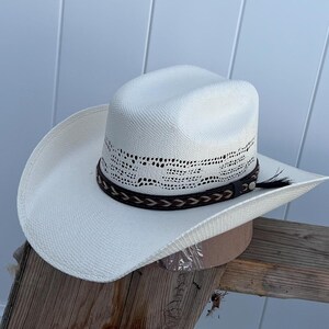 May include: A white straw cowboy hat with a brown leather band and a braided design. The hat has a wide brim and a tall crown.