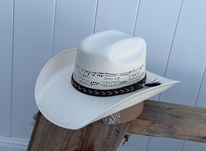 May include: A white straw cowboy hat with a brown leather band and a braided design. The hat has a wide brim and a tall crown.