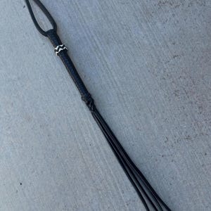 May include: A black leather riding crop with a white braided accent near the handle. The crop has a loop handle and a long, braided tail.