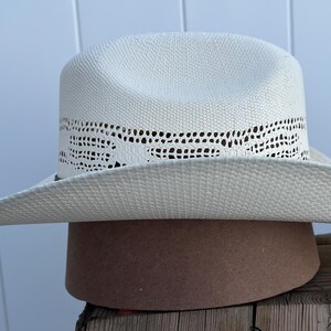 May include: A white straw cowboy hat with a woven design around the crown. The hat is sitting on a brown surface.