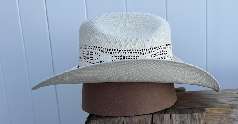 May include: A white straw cowboy hat with a woven design around the crown. The hat is sitting on a brown surface.