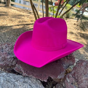 May include: A vibrant fuchsia cowboy hat with a classic design. The hat is made of a smooth material and features a structured crown and wide brim. It rests on a collection of dark, textured rocks, with a blurred outdoor background.