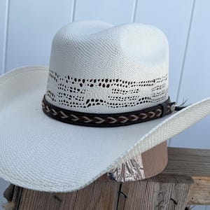 May include: A white straw cowboy hat with a brown leather band and a woven design. The hat has a wide brim and a tall crown.