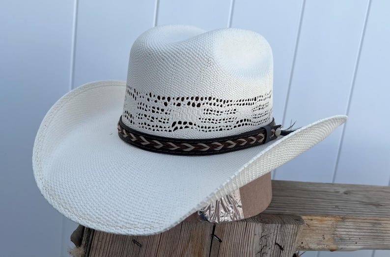 May include: A white straw cowboy hat with a brown leather band and a woven design. The hat has a wide brim and a tall crown.