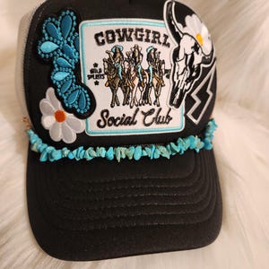 May include: Black trucker hat with a turquoise and white beaded trim. The hat features several patches, including a skull, a daisy, and a patch that says "Cowgirl Social Club" with a group of cowgirls riding horses.