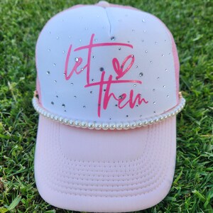 Puede incluir: Gorra de camionero rosa y blanca con ribete de perlas. La gorra tiene una parte trasera de malla blanca y una parte delantera rosa con las palabras "Let Them" en letras rosas con un símbolo de corazón.