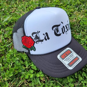 May include: A black and white trucker hat with a blue mesh back. The front of the hat has a red rose embroidered on it and the words "La Toxic" in black script.