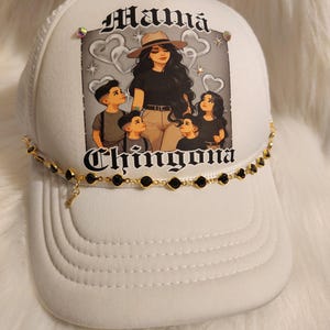 May include: White trucker hat with a black and white illustration of a woman and three children. The text "Mamá Chingona" is printed on the hat. The hat has a gold chain with black beads around the brim.