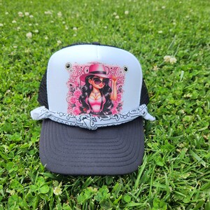 Könnte beinhalten: Schwarz-weiße Trucker-Cap mit weißem Frontpanel, das eine Grafik einer Frau mit rosa Hut und Sonnenbrille zeigt. Die Kappe hat einen schwarzen Schirm und ein Mesh-Rückenteil, mit weißem Bandana-Besatz.