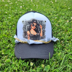 Könnte beinhalten: Schwarz-weiße Trucker-Cap mit einer Grafik einer Frau mit Sonnenbrille und Hut. Die Kappe hat eine goldene Kette und ein weißes Bandana. Die Kappe liegt auf grünem Gras.