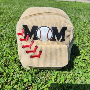 May include: A beige corduroy zippered pouch with the word "MOM" in black letters and a baseball graphic in the center. The pouch has red stitching around the edges.