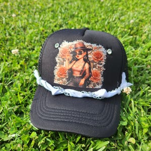 Könnte beinhalten: Schwarze Trucker-Mütze mit Mesh-Rückseite und gebogenem Schirm. Auf der Vorderseite befindet sich eine Grafik einer Frau mit Hut und Sonnenbrille, umgeben von Rosen. Die Mütze ist mit weißem Stoff und Strasssteinen verziert.