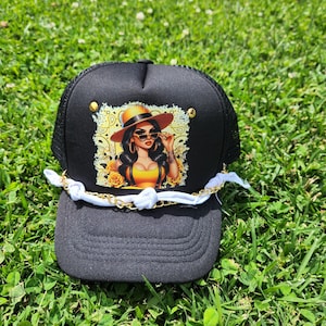 May include: Black trucker hat with a curved brim and mesh back. The front features a graphic of a woman wearing sunglasses and a hat. A white and gold chain is attached to the brim.