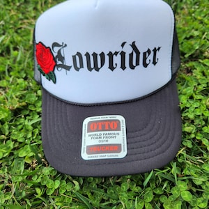 May include: A white and black trucker hat with a red rose and the word "Lowrider" in black lettering. The hat has a black mesh back and a black brim. The hat has a label that reads "OTTO World Famous Foam Front OSFM Trucker Durable Snap Closure".