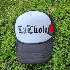 May include: A white and black trucker hat with a black embroidered design that says "La Chola" and a red rose patch on the side.