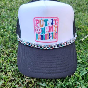 May include: White and black trucker hat with a colourful embroidered patch that says "PUT IT ON MY HUSBANDS TAB". The hat has a silver chain around the brim.