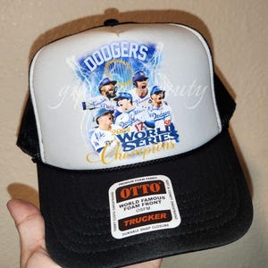 May include: A black and white trucker hat with a white front panel featuring a graphic of the Los Angeles Dodgers baseball team celebrating their 2020 World Series Championship. The text "DODGERS 2020 World Series Champions" is printed on the hat. The hat has a black mesh back and a black adjustable snap closure.