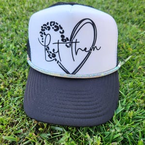 Puede incluir: Una gorra de camionero blanca con un diseño de corazón negro y las palabras "Let Them" dentro del corazón. El corazón tiene un estampado de leopardo. La gorra tiene una visera negra y un ribete de purpurina plateado.