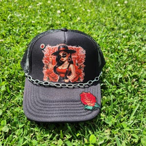Könnte beinhalten: Schwarze und graue Trucker-Mütze mit Mesh-Rückseite und dekorativer Kette. Die Vorderseite zeigt eine Grafik einer Frau mit Hut und Sonnenbrille, umgeben von Rosen. Ein roter Rosenaufnäher befindet sich auf dem Rand.