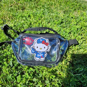 Puede incluir: Riñonera transparente con ribete y correa negros. La parte delantera presenta un parche de Hello Kitty con una gorra y camiseta de béisbol de los Dodgers, sosteniendo una taza. También hay un gráfico de sol rojo en la parte delantera.