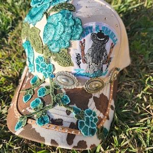 Puede incluir: Gorra de béisbol beige con visera marrón con estampado de vaca. La gorra está adornada con bordados florales turquesa, adornos plateados y un parche con un gráfico de vaquera. Tiene un botón marrón en la parte superior, sobre hierba verde.