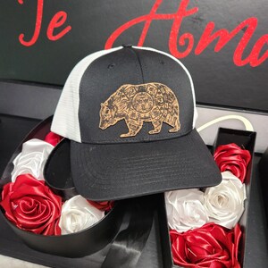May include: A black and white trucker hat with a brown leather bear patch. The bear is surrounded by a circle of symbols. The hat is sitting on top of a black box filled with red and white silk roses.