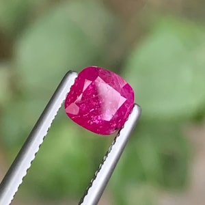 Könnte beinhalten: Ein leuchtend roter Rubin im Kissenschliff, gehalten von einer silbernen Pinzette. Der Edelstein zeigt eine tiefe, gesättigte rote Farbe mit sichtbaren Facetten. Der Hintergrund ist unscharf, mit grünem Laub.