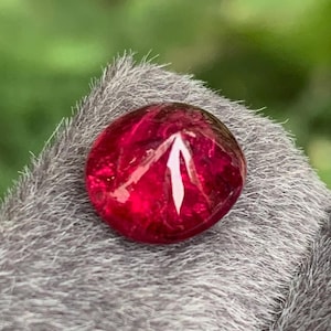 May include: A circular, polished, deep red gemstone with a bright, reflective surface. The gem is set against a soft, grey background, with a blurred green backdrop. The gemstone's colour is a vibrant, saturated red.