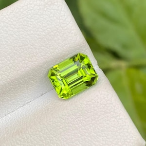 Könnte beinhalten: Ein leuchtend grüner, rechteckig geschliffener Peridot-Edelstein. Der Edelstein liegt auf einer weißen Oberfläche, mit einem verschwommenen grünen Hintergrund. Die Facetten des Steins reflektieren das Licht und zeigen seine Klarheit und Brillanz.