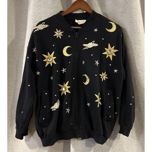 May include: Black zip-up jacket with celestial-themed embellishments. Features gold stars, crescent moons, and rhinestone accents. The jacket has a relaxed fit with pockets and a ribbed collar and cuffs. A unique and stylish outerwear piece.