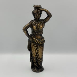 May include: Bronze-colored statue of a woman in classical attire, holding a vessel on her head. The figure is draped in flowing robes and has one arm raised. The statue is approximately 10 inches tall and stands on a flat base.