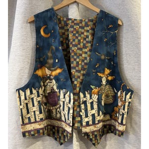 May include: Reversible Halloween vest with a navy blue background and a whimsical design. One side features a witch and scarecrow with a fence, the other side has a patchwork pattern. The vest is hanging on a wooden hanger.