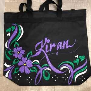 May include: Black tote bag with a purple and green floral design and the name "Kiran" written in purple cursive.