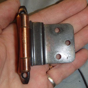 May include: A cabinet hinge with a copper-colored barrel and a silver-colored mounting plate. The plate has three holes for screws and the text "PAT. 1967". The hinge is designed for use on cabinets and doors.