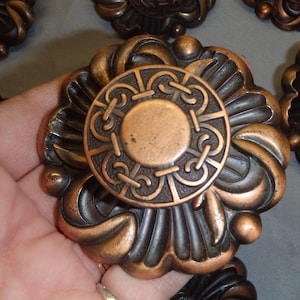 May include: Ornate copper-colored cabinet knobs with a floral design. Each knob features a central circular medallion with an intricate pattern. The knobs have a weathered, antique appearance, with detailed relief work and a warm, metallic finish.