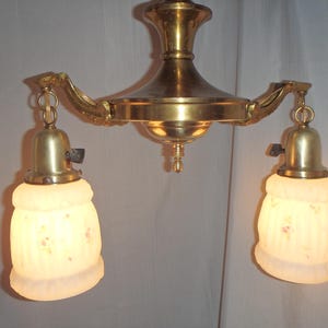 May include: Antique brass chandelier with two frosted glass shades. The shades are decorated with small pink floral patterns. The fixture has a classic design, with ornate arms and a central support. The light fixture is designed to be mounted on a ceiling.
