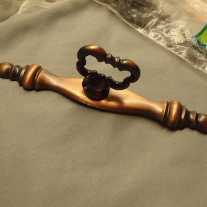 May include: A bronze-toned cabinet pull with a decorative handle. The pull has a central handle with an ornate design and is attached to a rectangular base with rounded ends. The hardware is new.