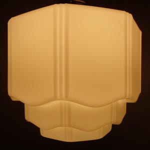 May include: A vintage, off-white glass pendant light fixture. The light has a geometric, layered design with a rectangular top and a scalloped bottom. Vertical lines add detail to the sides. The fixture is suspended from a dark ceiling.