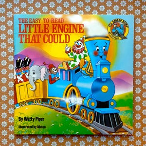 Könnte beinhalten: Ein Vintage-Kinderbuchcover mit einer blauen Lokomotive mit einem lächelnden Gesicht. Der Zug zieht einen gelben Güterwagen, auf dem ein Clown, ein Elefant und ein Affe sitzen. Der Titel des Buches lautet "The Easy-to-Read Little Engine That Could".