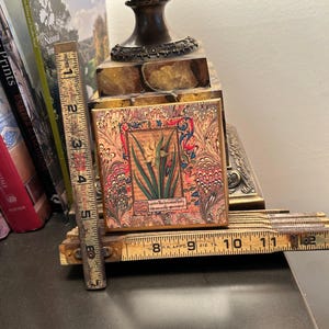 May include: A small, ornate wooden box with a floral design. The box is closed and has a gold-colored frame. The box is sitting on a wooden ruler.