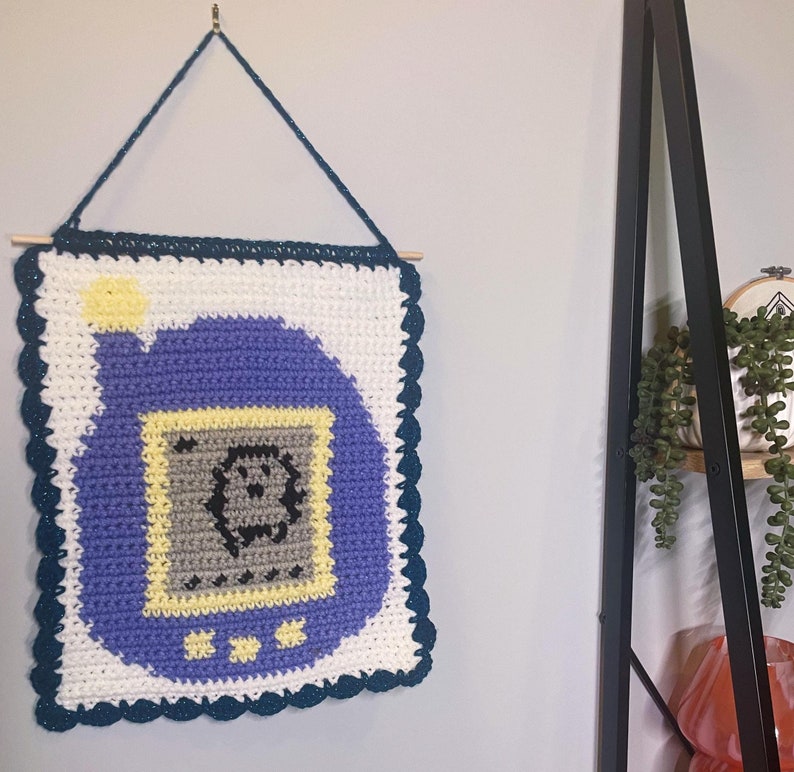 May include: A crocheted wall hanging featuring a purple and white pixelated design of a handheld digital pet, similar to a Tamagotchi.