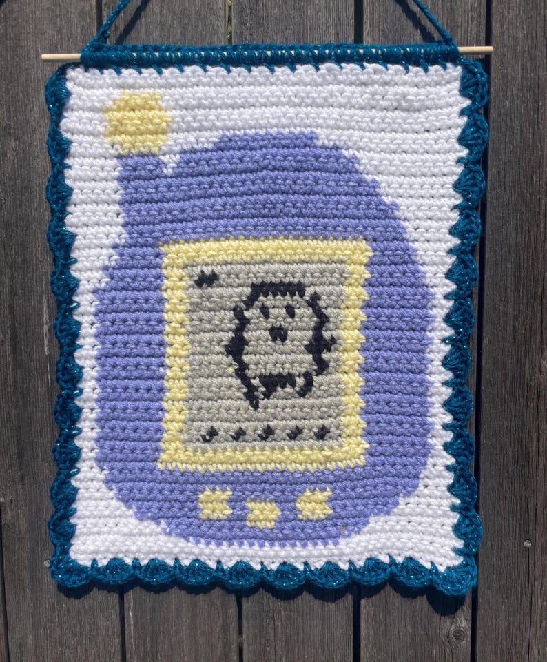 May include: A crocheted wall hanging depicting a pixelated image of a purple and yellow Tamagotchi, a popular virtual pet toy. The hanging is framed with a white border and a teal scalloped edge.