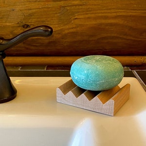 May include: A teal soap bar sits on a wooden soap dish. The dish has a decorative, stepped design. The soap dish is on a white porcelain sink.