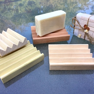 May include: Several wooden soap dishes in various designs. One dish is holding a bar of soap. The soap dishes are made of light-colored wood and have a slatted design to allow for drainage. One soap dish is made of a darker wood.