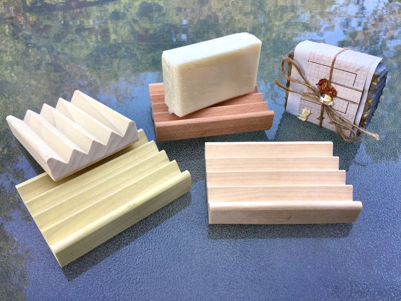 May include: Several wooden soap dishes in various designs. One dish is holding a bar of soap. The soap dishes are made of light-colored wood and have a slatted design to allow for drainage. One soap dish is made of a darker wood.