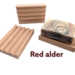 May include: Three red alder wood soap dishes. One dish holds a bar of soap with a marbled design in shades of red, blue, and white. The other two dishes are empty. The text "Red alder" is visible.
