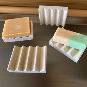 May include: Several wooden soap dishes with a grooved design are displayed, some holding bars of soap. The soaps vary in color, including shades of tan and green. The dishes are made of light-colored wood.